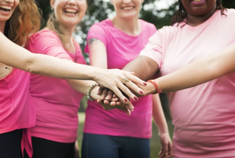 grupo de mulheres diversas usando camisa rosa e dando as mãos representando o Outubro Rosa no ambiente de trabalho