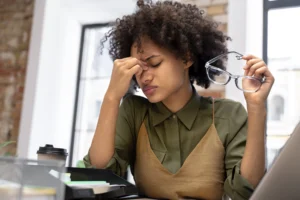 mulher negra de cabelos crespos e volumosos com a mão nos olhos aparentando estar estressada no local de trabalho - nova nr1