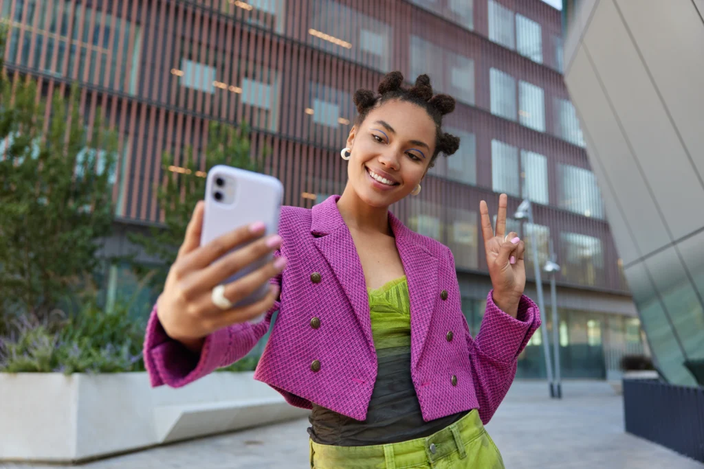 attractive girl fashionable outfit creats influence content shows peace sign smartphone front camera poses selfie smiles gladfully poses urban place spends vacation big city employee advocacy